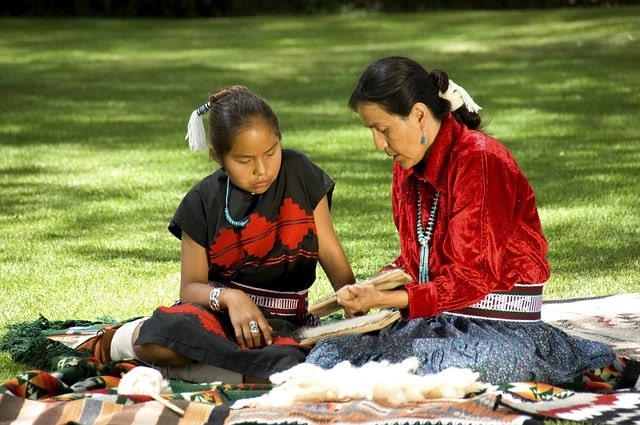 an indigenous grandmother and granddaughter sitting on the ground working on a craft