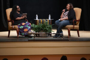 two women sit in chairs on a stage