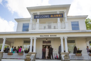 people gather on the porch of an old house with columns and a banner that reads 