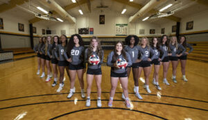 The Brenau volleyball team stands in a triangular formation in front of the net. The player in front each hold a volleyball.