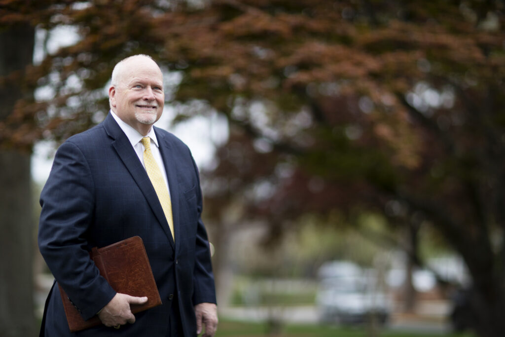 Brenau President David L. Barnett, Ph.D. holds a folio and smile against a blurred background of trees